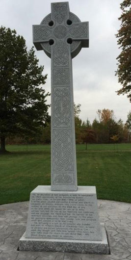 Large grey Celtic Cross with ornate patterning and inscription on plinth. The cross is positioned on a grey circular dais, with green grass and trees visible in background.