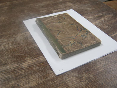 Brown diary with fraying green binding, lying on white paper, on brown table with scuffed surface.