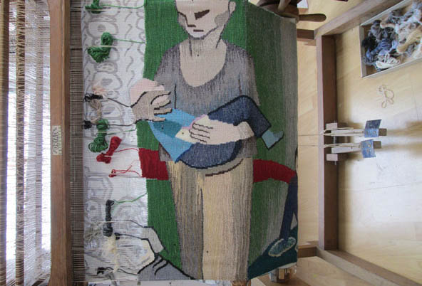 Image of partially completed tapestry being woven on a loom. It features a stylized, cartoonish figure of a woman in a grey top and light trousers carrying a child close to her chest and walking forward, against a green backdrop with a red stripe. Numerous spools of yarn and artist’s studio visible in background.