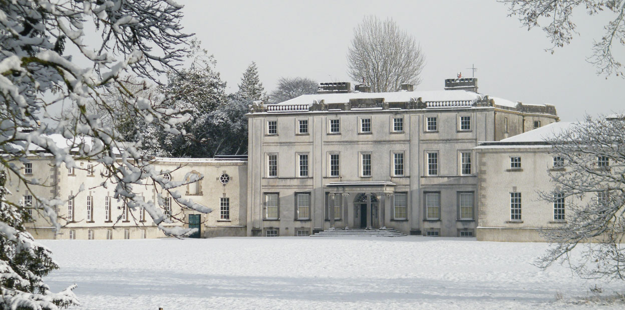 Une grande bâtisse blanche de trois étages et munie de deux pavillons, aperçue en façade, pendant l’hiver. La neige recouvre le sol et plusieurs arbres sont visibles.