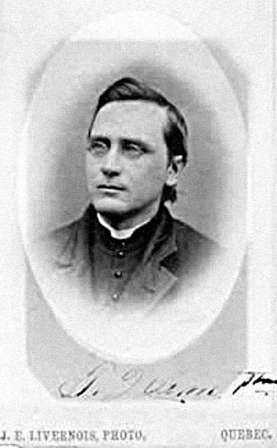 Black and white upper portrait of man wearing glasses with dark hair. He is wearing a dark jacket, vest, and  a priest’s collar. T. Quinn. Priest handwritten at bottom of photograph. J.E. Livernois. Photo. Quebec printed below photograph.